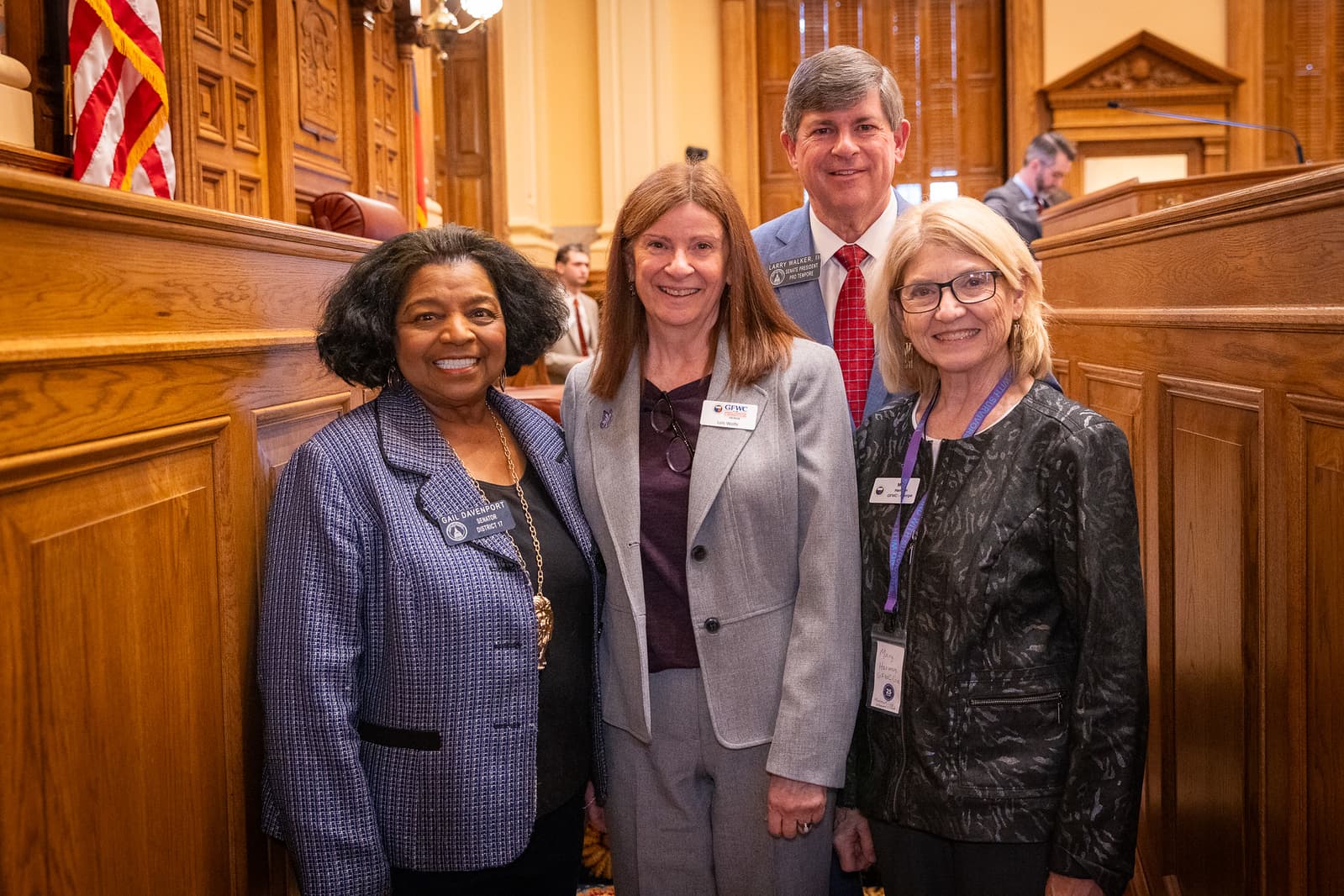 Four smiling professionals pose together in a formal, wood-paneled legislative chamber with flags.