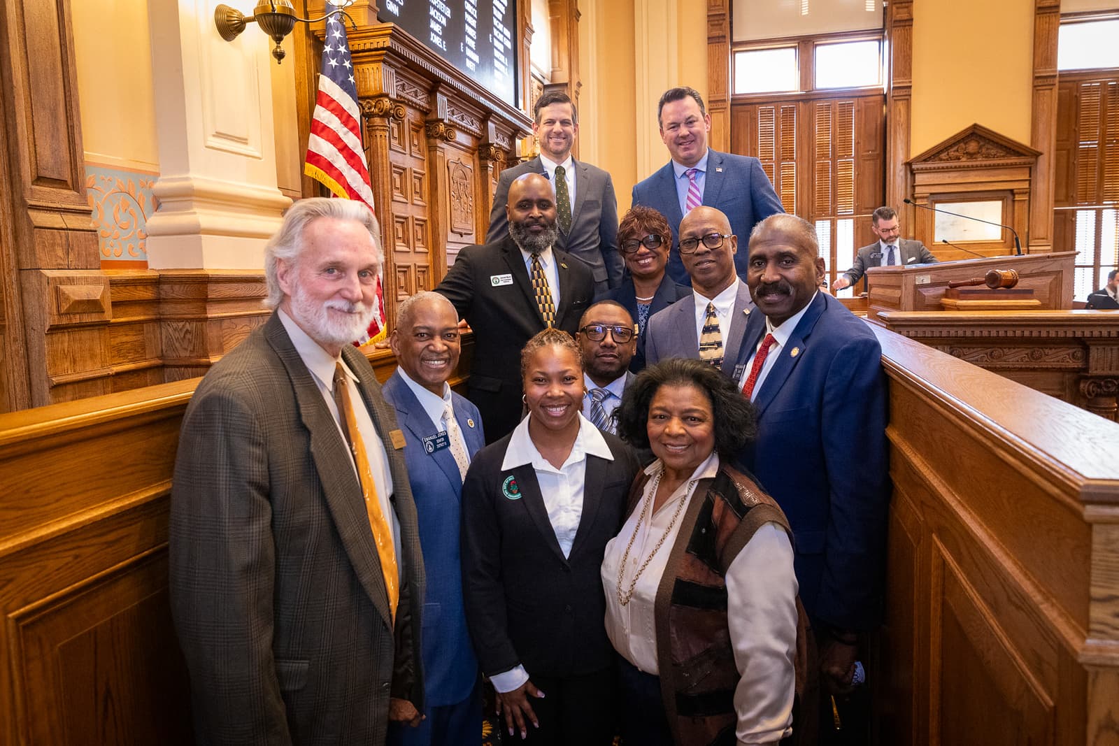 Diverse group of professionals in business attire posing inside a wood-paneled government legislative chamber.