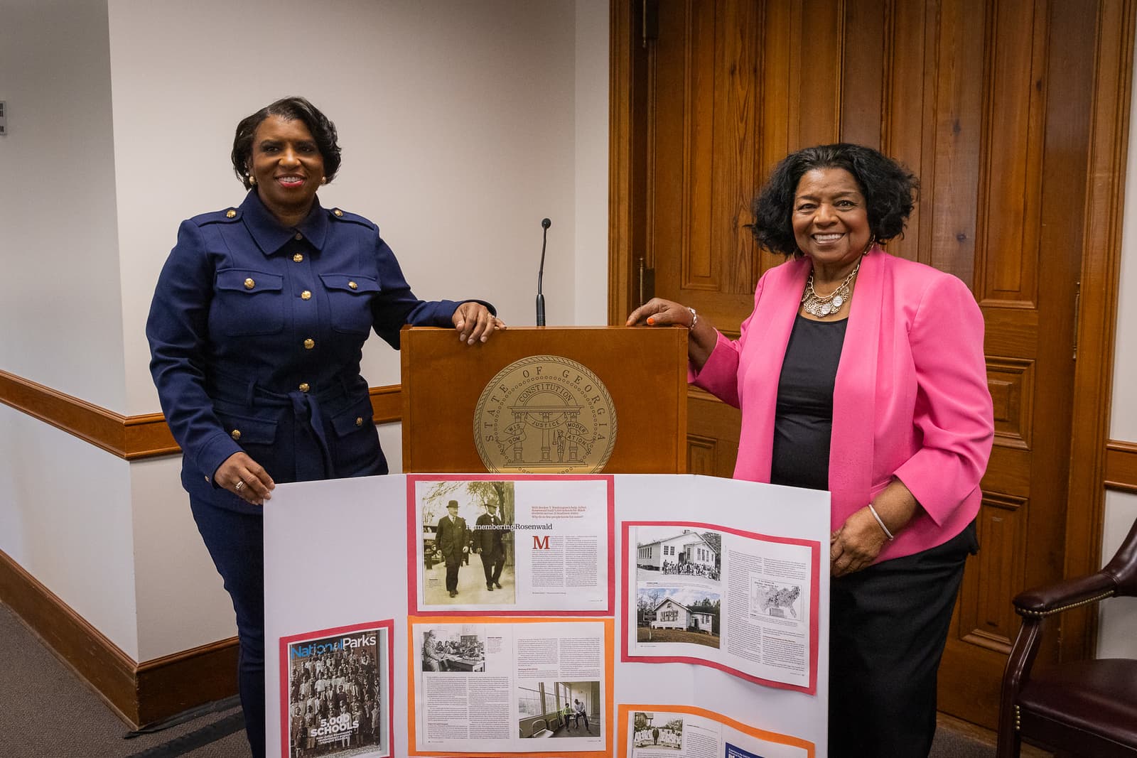 Two women stand behind a State of Georgia podium and a Rosenwald schools display board.