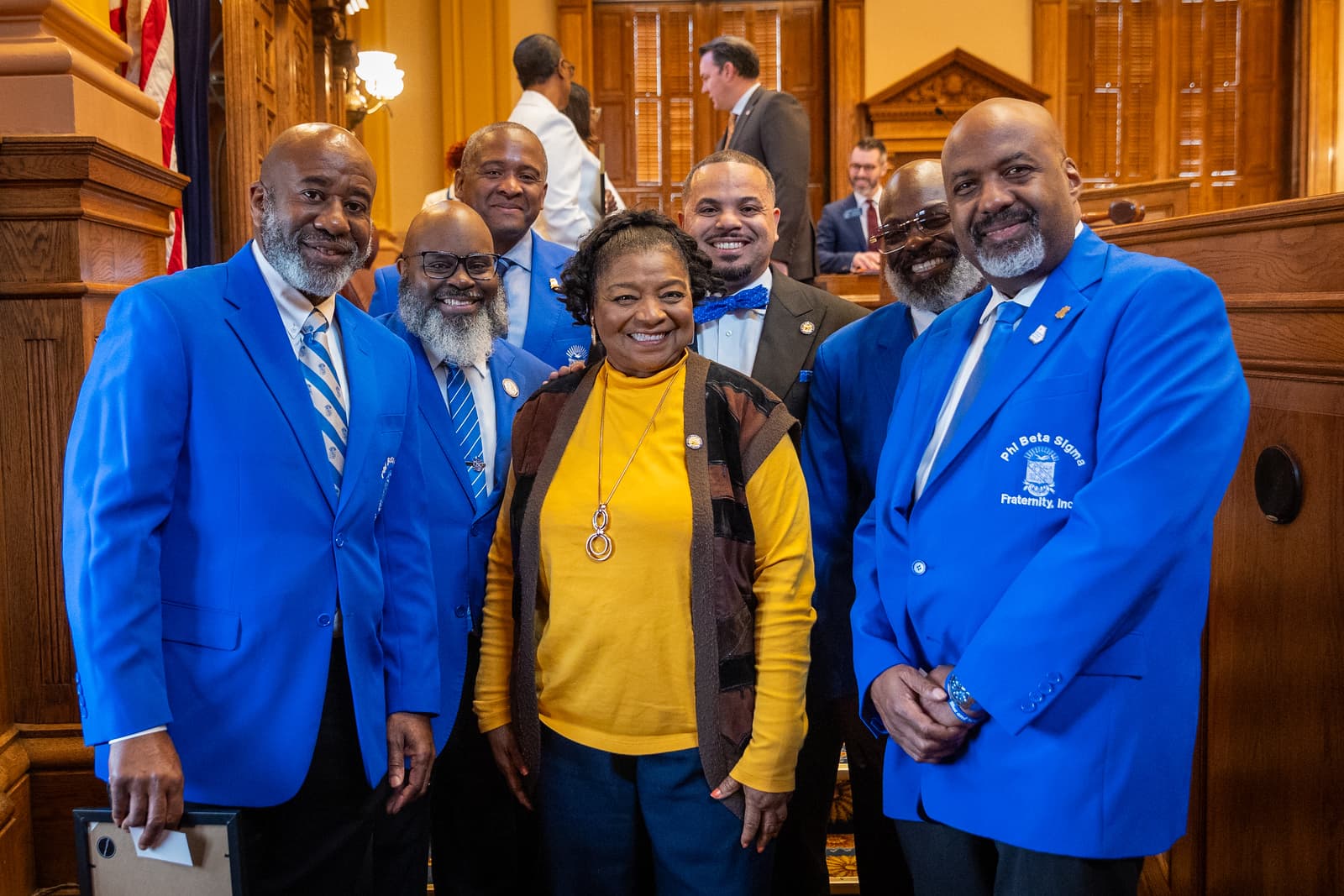 Phi Beta Sigma members in blue blazers pose with a woman in a yellow top.