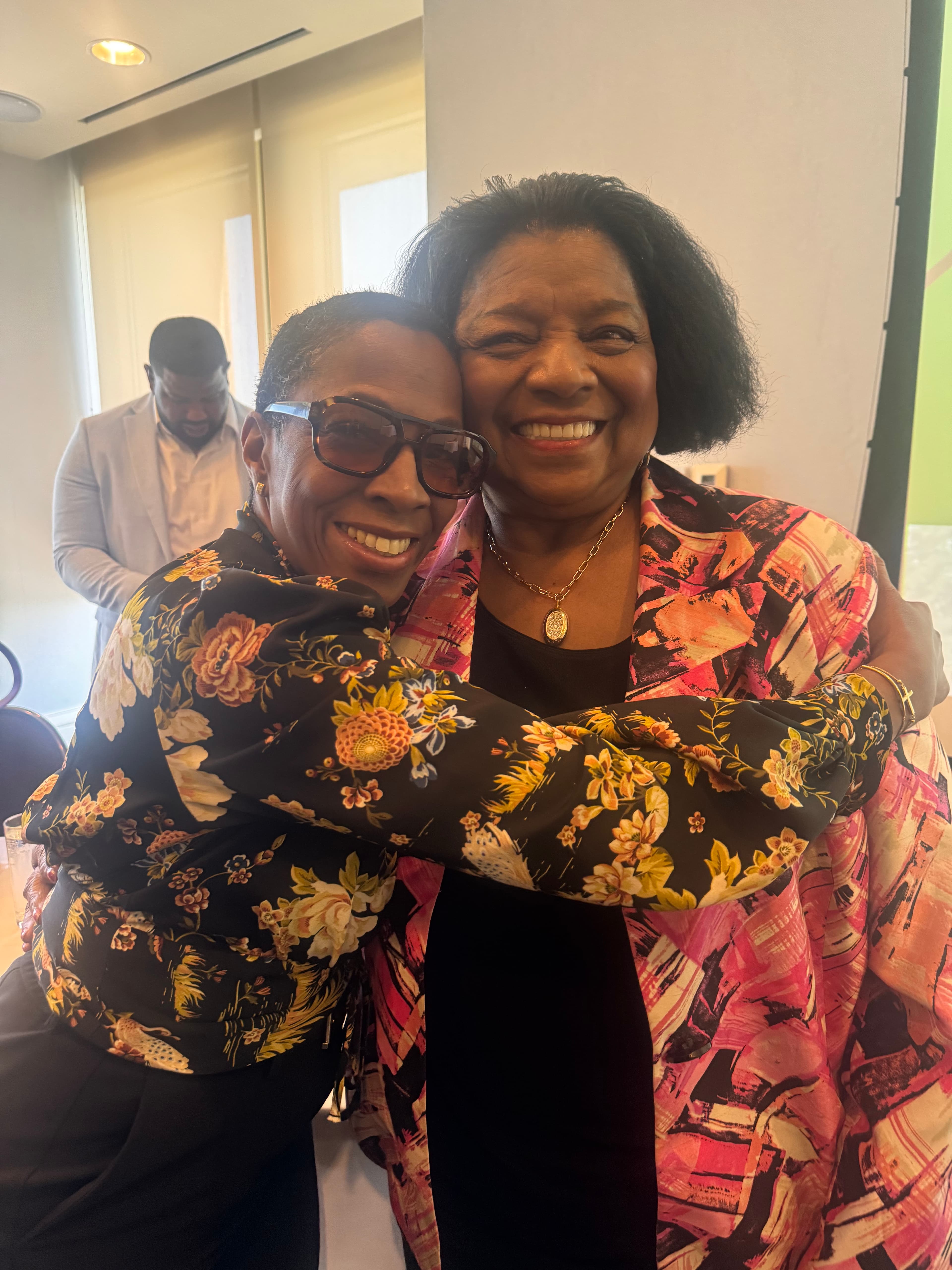 Two joyful Black women hugging, wearing a pink patterned jacket and a floral blouse.